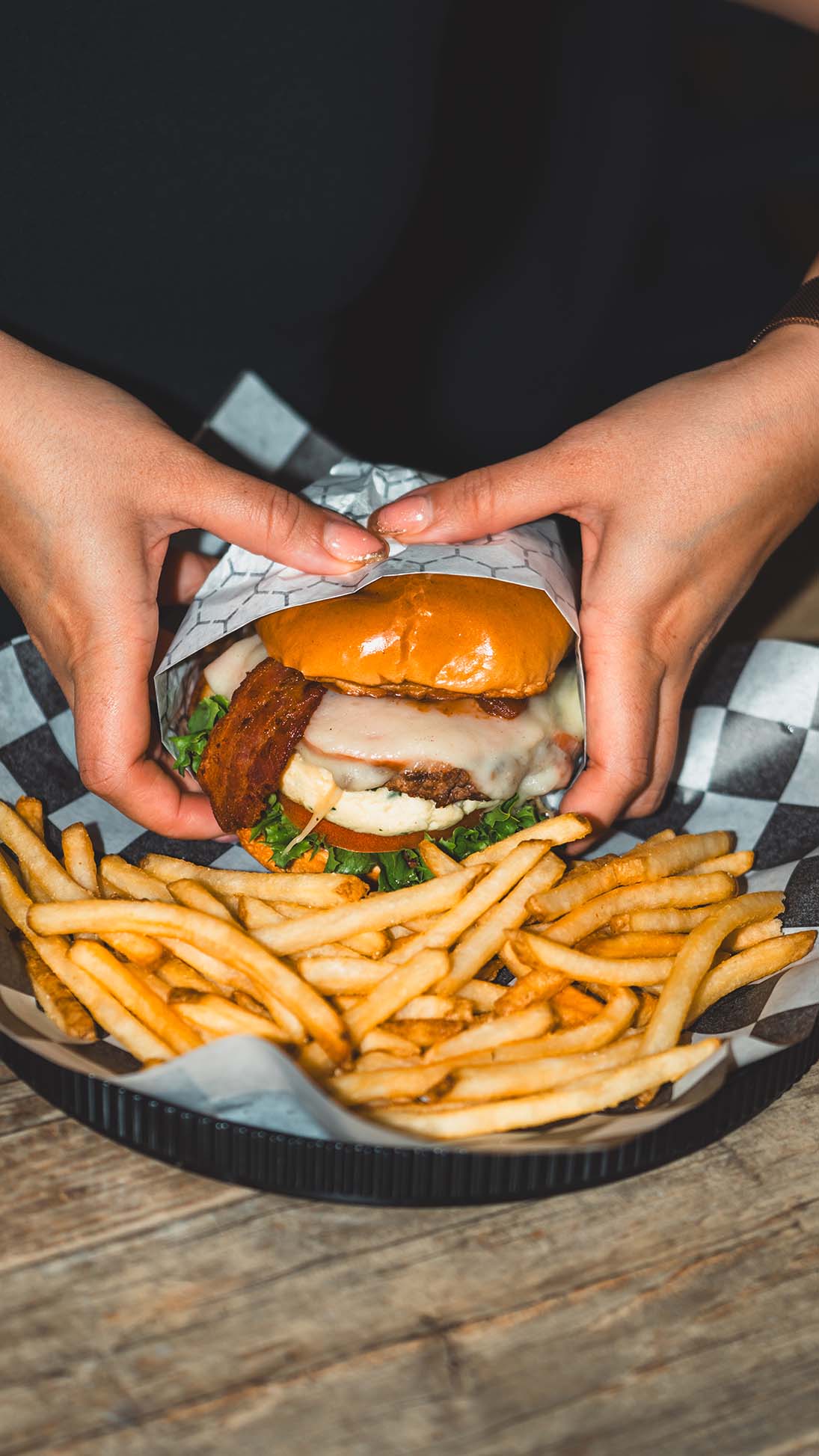 Hands framing a bacon cheeseburger above a generous portion of fries.