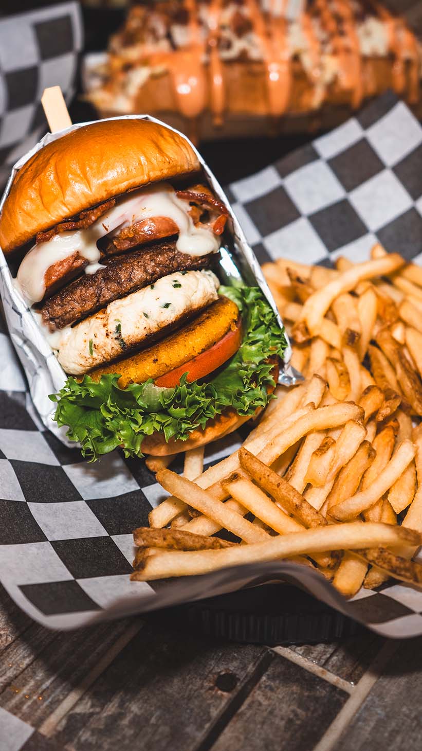 Towering specialty burger and fries arranged on checkered deli paper.