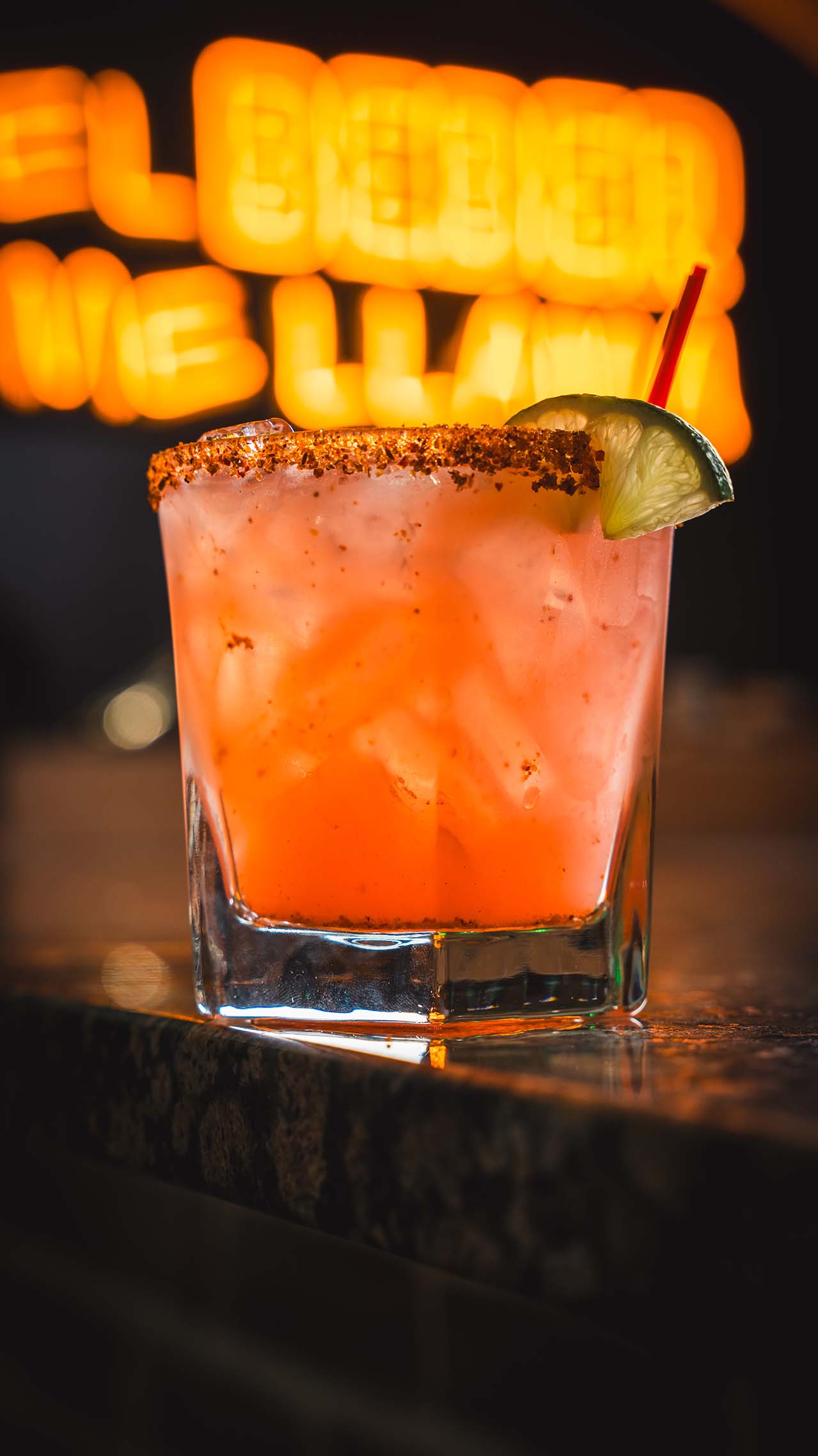 Pink cocktail with spiced rim and lime against glowing restaurant signage.