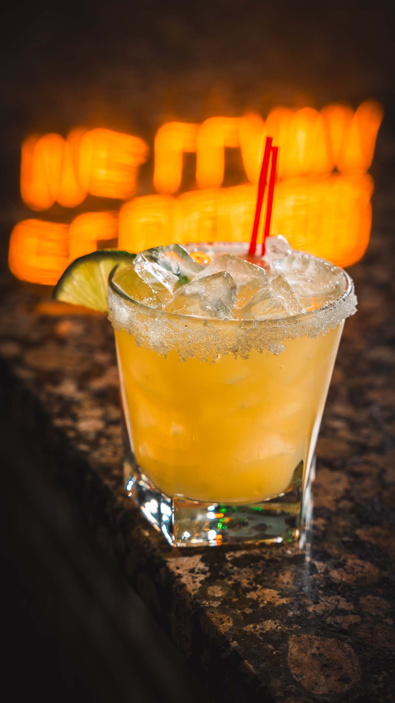 Golden margarita with sugar rim and lime against warm neon signage.