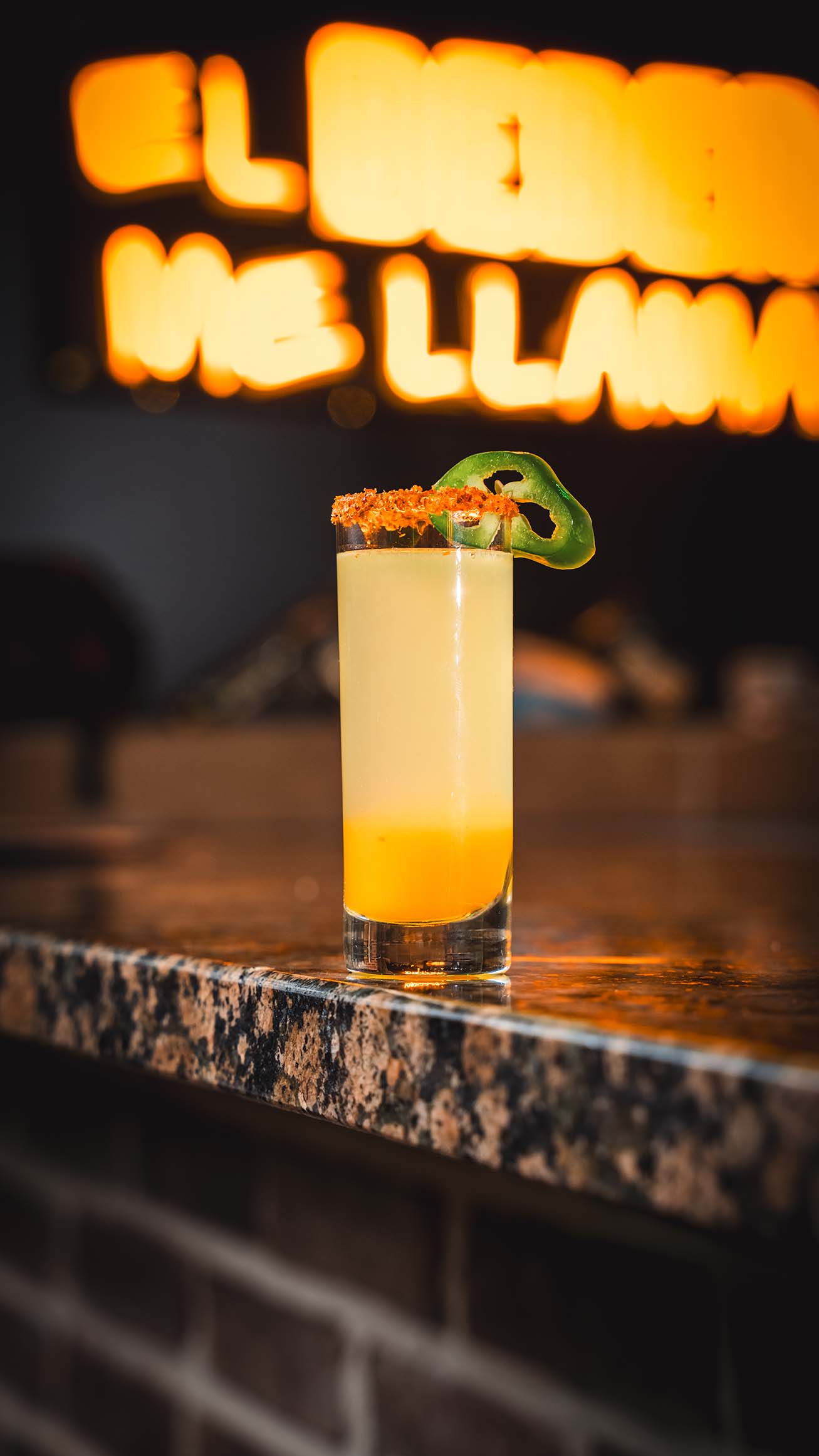 Tall citrus cocktail garnished with jalapeño against glowing bar signage.