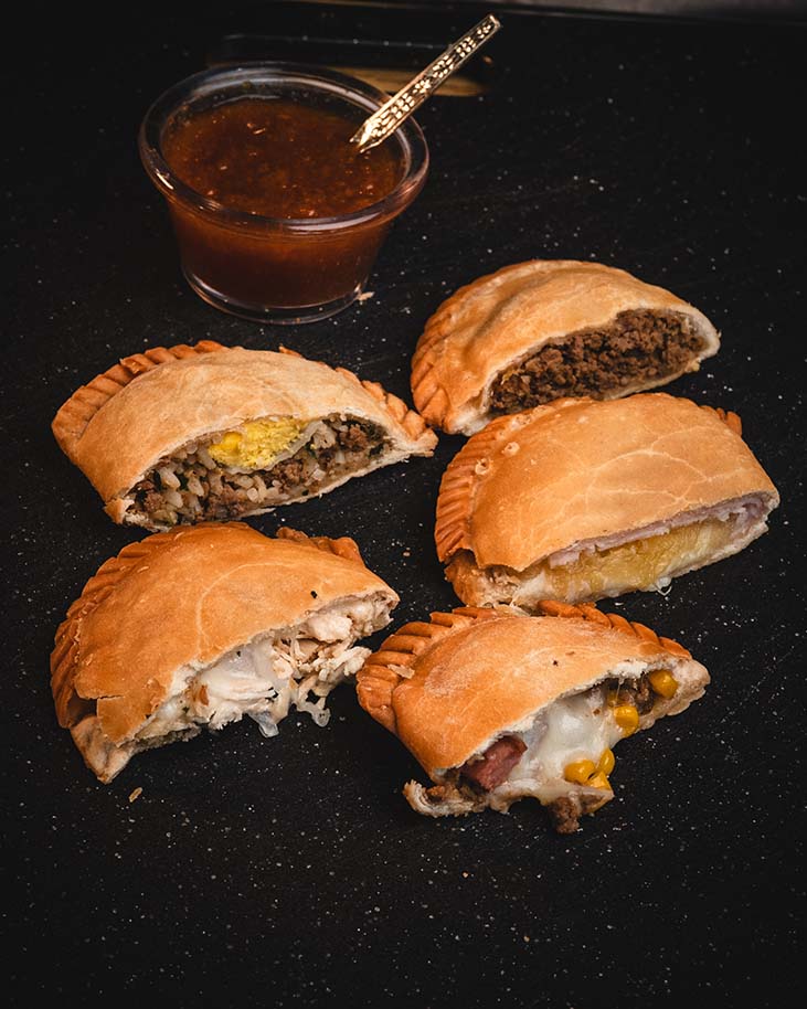 Assorted handmade empanadas with dipping sauce on a dark surface.