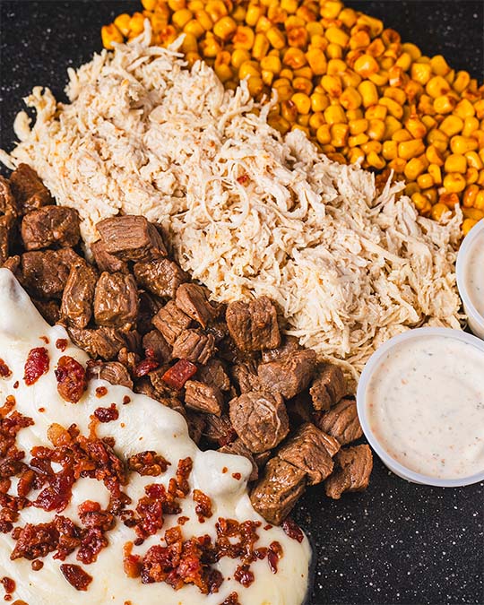 Close food composition of shredded chicken, grilled beef, corn, and creamy sauces.
