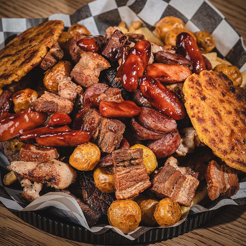 Abundant mixed-grill platter with sausages, pork, beef, potatoes, and arepas.